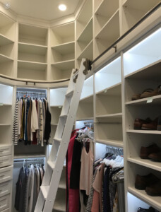 A walk-in closet with white shelves, hanging clothes, shoes, and an attached rolling ladder for access to upper storage areas.