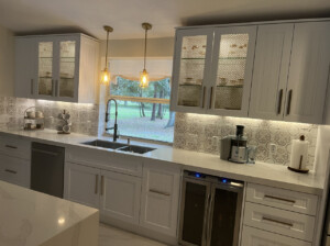 Modern kitchen with white cabinets, a large window, and patterned backsplash. Includes stainless steel sink, appliances, and under-cabinet lighting. Two pendant lights hang above the countertop.