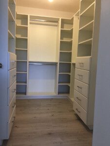 A walk-in closet with wooden flooring, multiple shelves, drawers, and two hanging rods. The closet is empty and well-lit.