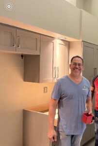 A man in a blue t-shirt stands in a kitchen near a counter, smiling and holding a red object.