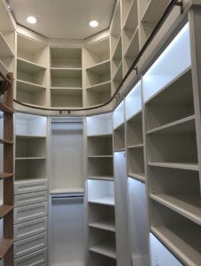 A walk-in closet with white shelving units, drawers, and hanging rods. Shelving extends from floor to ceiling, with a rolling ladder on the left side for access to higher shelves. Ceiling lights illuminate the space.