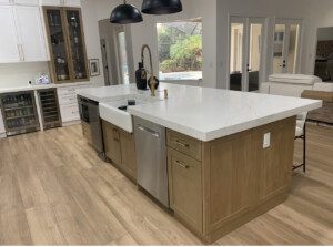 Modern kitchen with a large white marble island, wooden cabinets, stainless steel appliances, and black pendant lights. The kitchen has a clean, minimalist design with ample natural light.