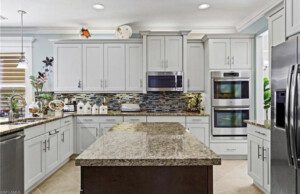 A modern kitchen with white cabinets, granite countertops, an island, stainless steel appliances, and a mosaic tile backsplash. Decor includes plants and kitchen canisters.