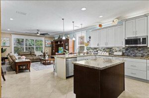 Modern kitchen and living area with white cabinets, granite countertops, a center island, sofa, coffee table, and a ceiling fan. Decor includes hanging plants and wall art. Tile flooring throughout.