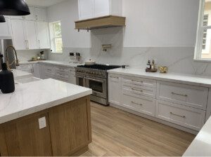 A modern kitchen with white cabinets, marble countertops, stainless steel appliances, and wooden accents. The floor is wooden, and the sink area includes a black faucet and decor items.