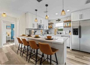 Modern kitchen with a central island, four amber-colored stools, stainless steel appliances, and white cabinetry. The backsplash features gray tiles, and pendant lights hang over the island.