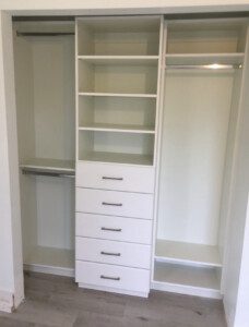 A white built-in closet with multiple shelves, drawers, and hanging rods, set against a light-colored wall on a gray wooden floor.